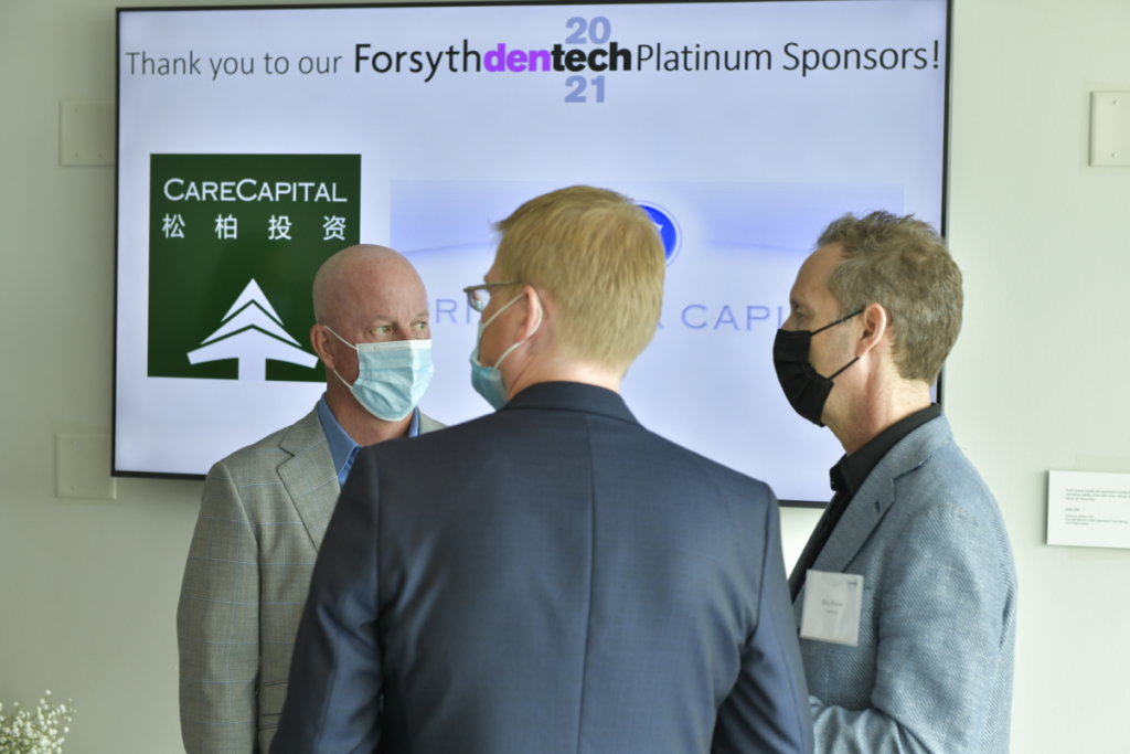 3 white male participants of dentech 2021 standing in front of screen with platinum sponsors' logos featured.
